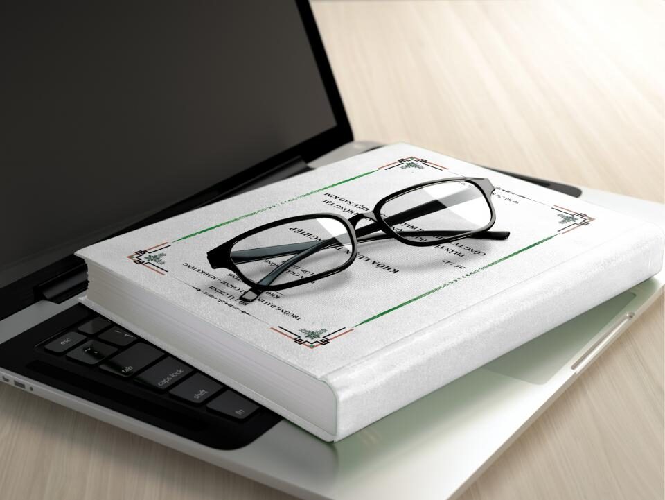 A pair of reading glasses resting on a closed book beside a laptop