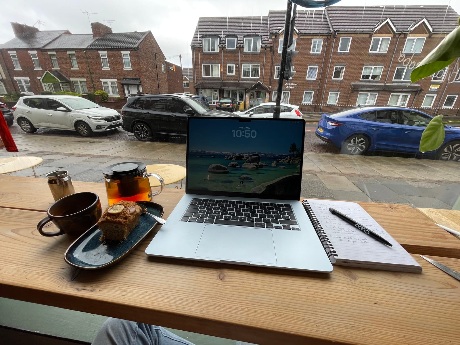 A laptop, a notebook and a pot of tea on a café window bench, looking out on a wet street